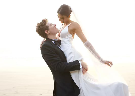 Man in a suit lifting a woman in a white dress against a bright background