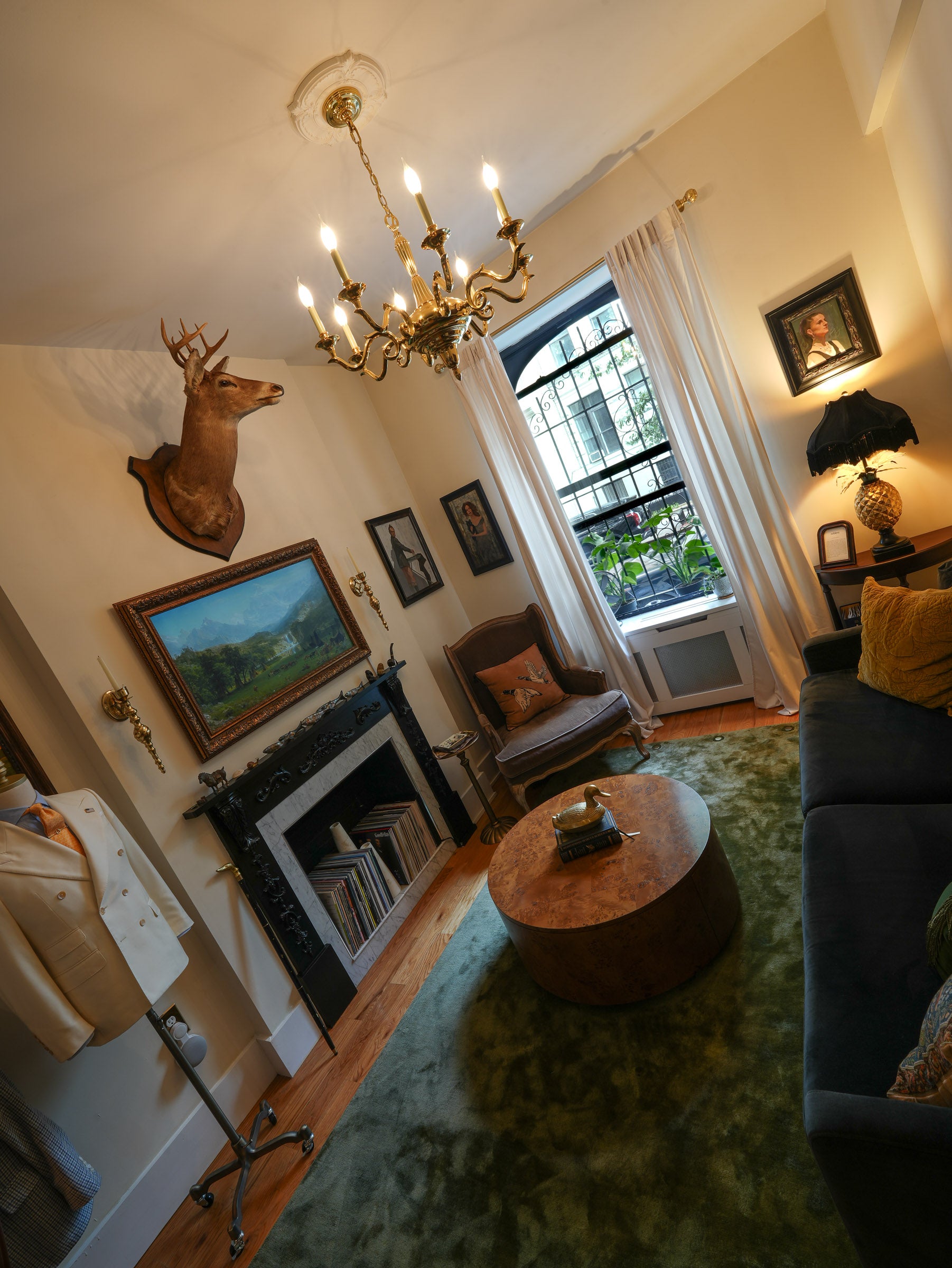 A cozy, vintage-style living room with warm cream walls, a gold chandelier, a fireplace with framed art, a green patterned rug, a round wooden coffee table, and a dark blue sofa with yellow pillows.