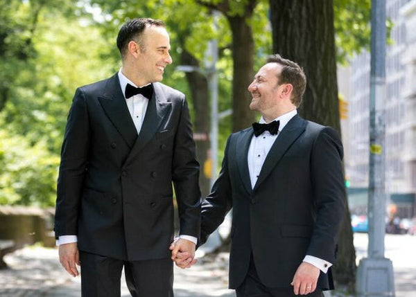 Two men in formal suits holding hands outdoors with trees and buildings in the background.