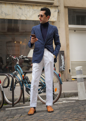 Man in a blue pinstripe blazer and white pants standing on a street, looking at his phone.
