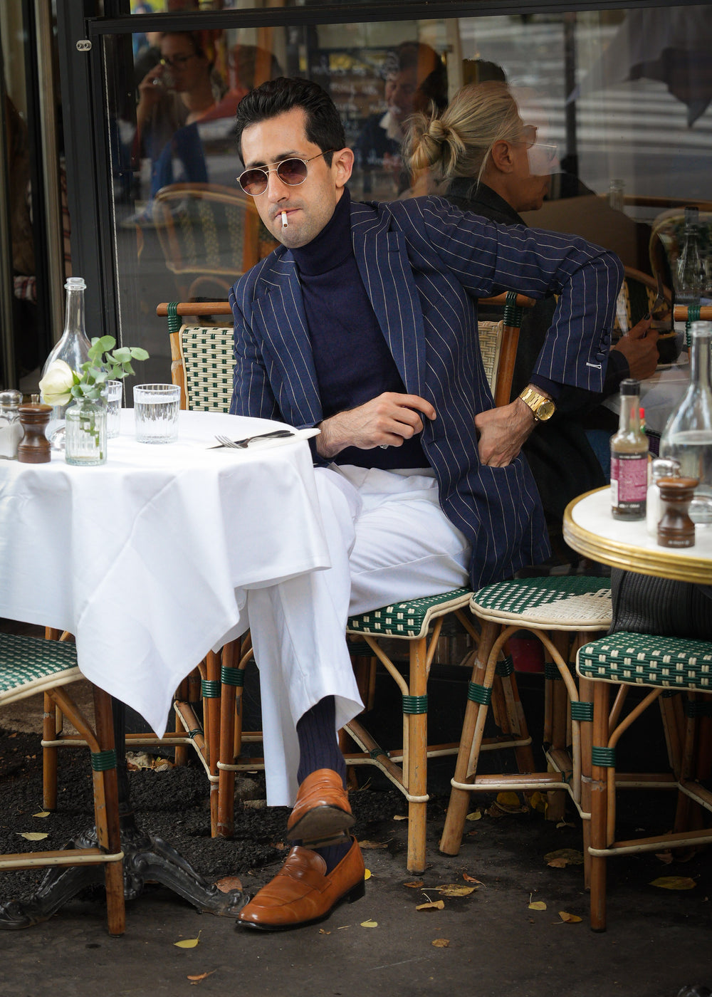 Man in a blue pinstripe blazer and white pants standing on a street, looking at his phone.