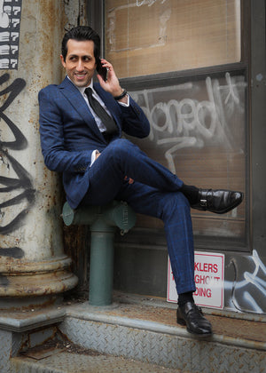 Man in a blue suit sitting on a pipe, talking on a phone, with graffiti in the background.