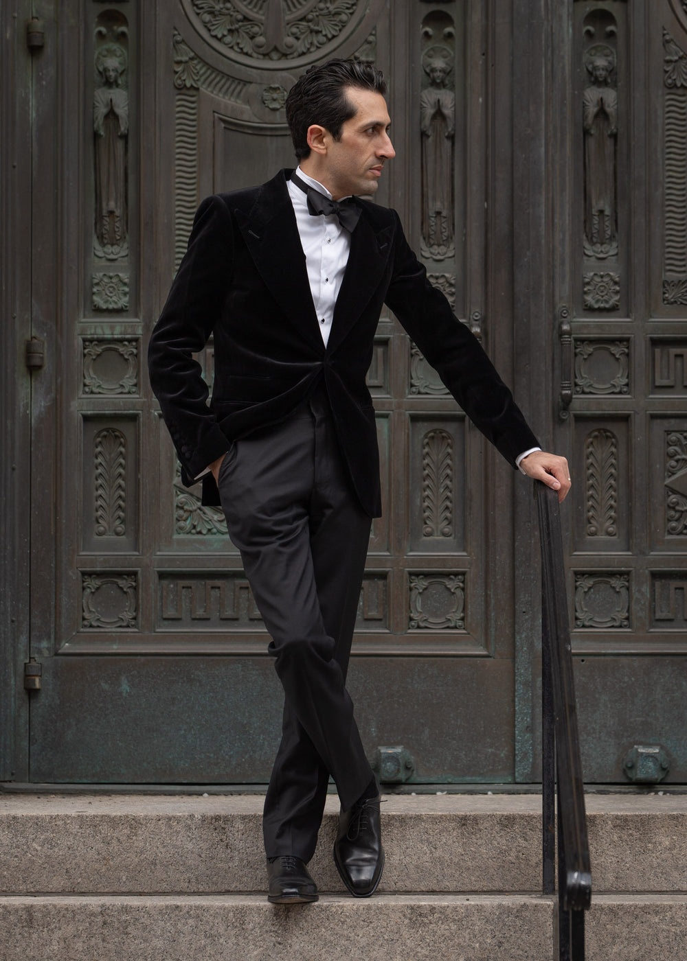 Man in a black suit standing on steps in front of an ornate door.
