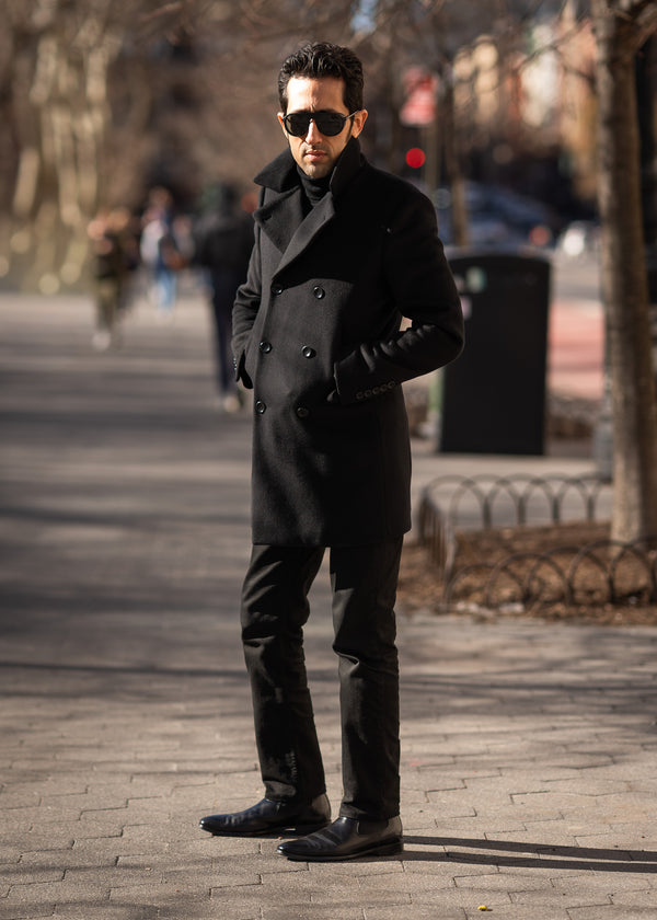 Man in a black coat and sunglasses standing on a paved walkway.