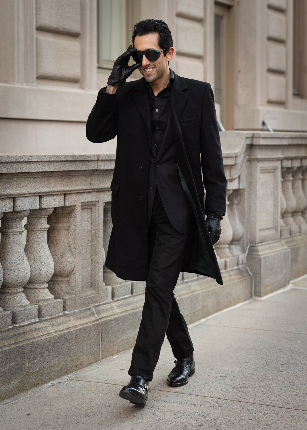 Man in a black coat and suit walking on a sidewalk with a building in the background