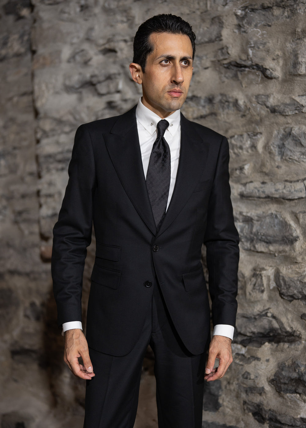 Man wearing a black suit and tie against a stone wall.