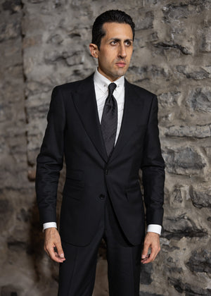 Man wearing a black suit and tie against a stone wall.