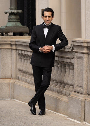 Man in a black tuxedo standing against a stone building.