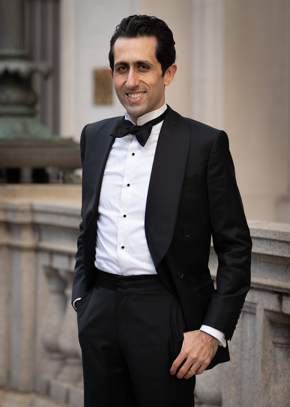 Man in a black tuxedo standing outdoors with classical architecture in the background