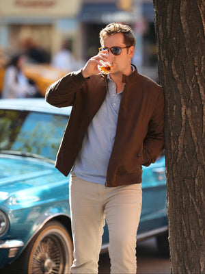 A man in a brown suede bomber jacket drinking a glass of whiskey in front of a vintage blue car.