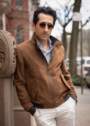 Man wearing a brown leather jacket and sunglasses leaning against a wall outdoors.