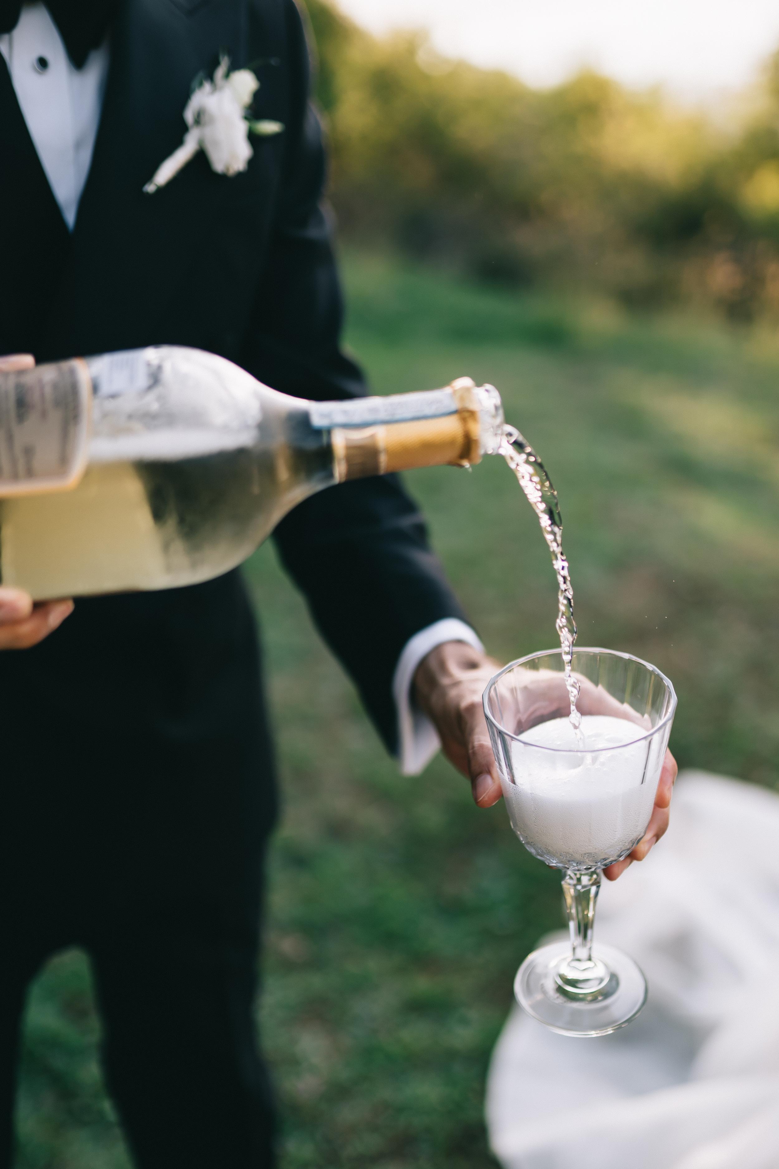 Person in a tuxedo pouring champagne into a glass outdoors.
