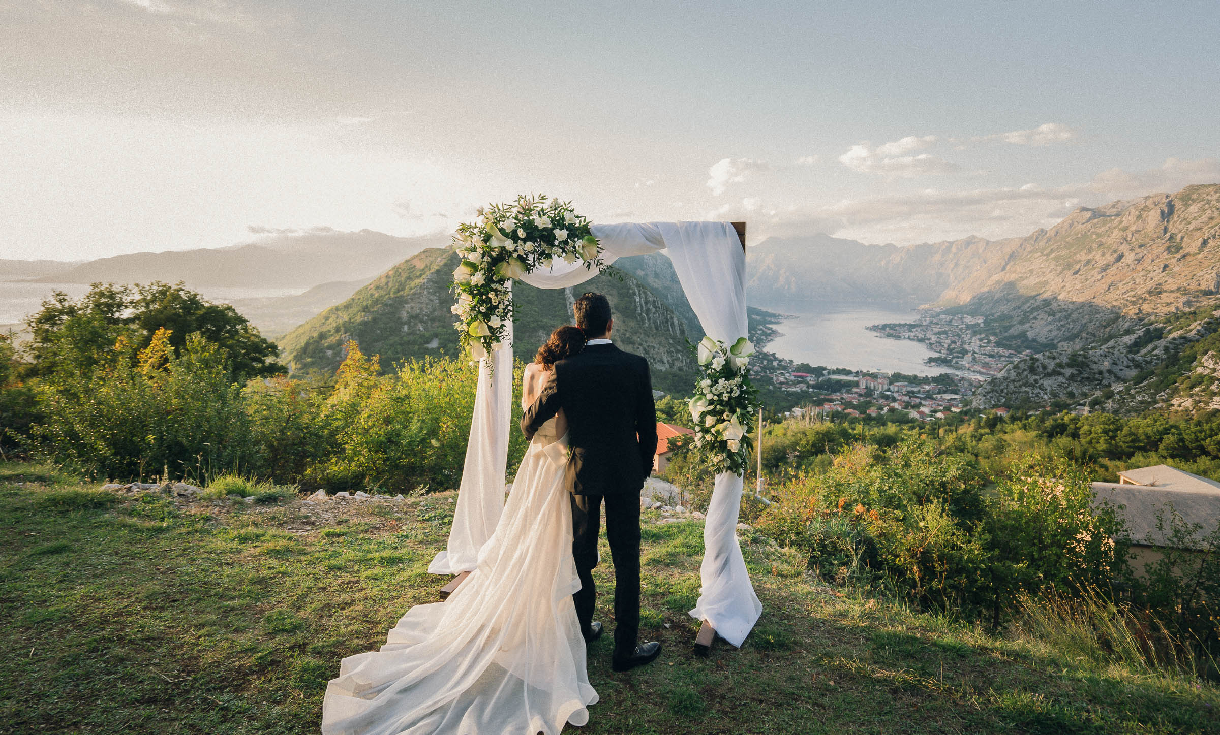 Couple standing under a wedding arch with a scenic view of mountains and water.
