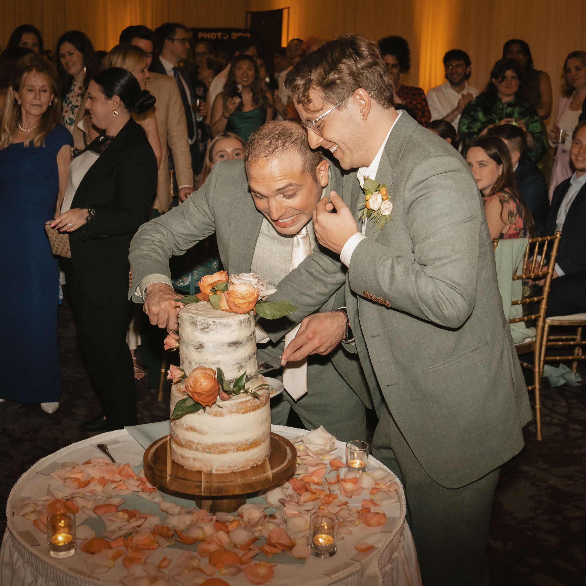 Two men in suits cutting a wedding cake with guests in the background