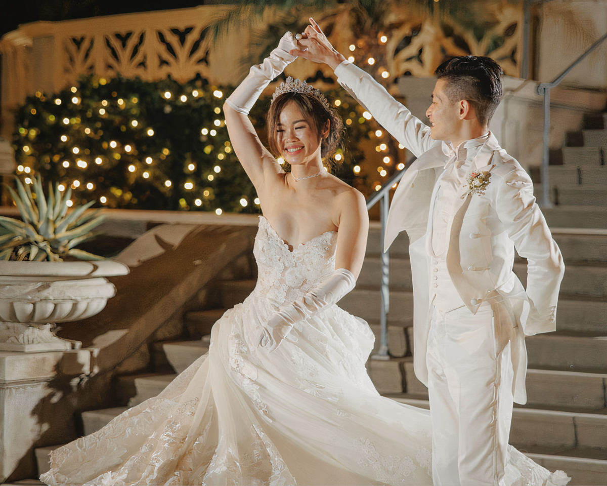 Couple in wedding attire dancing outdoors with decorative lights and plants in the background.