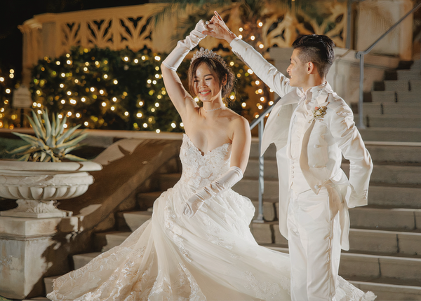 Couple in wedding attire dancing outdoors with decorative lights and plants in the background.