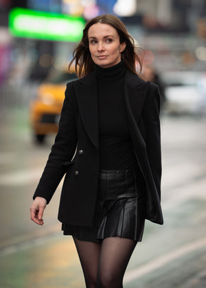 Woman in a black coat and leather skirt walking on a city street with blurred background