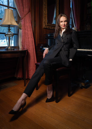 Woman in a black suit sitting on a piano in a room with red curtains and a lamp.