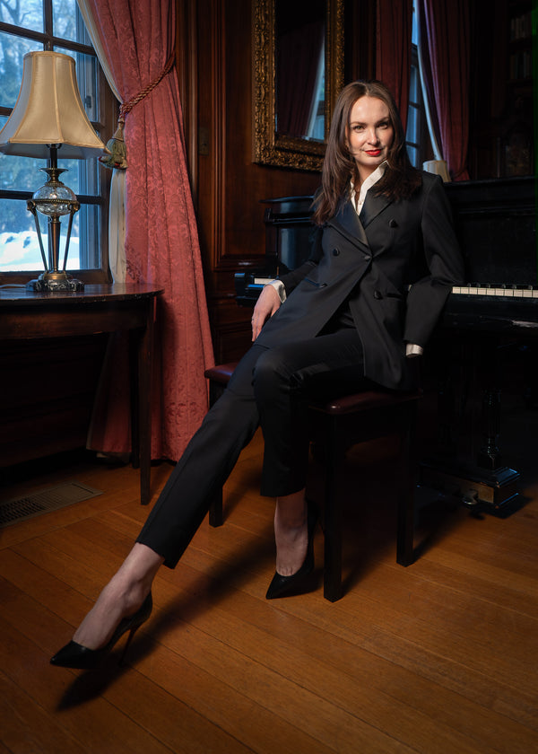 Woman in a black suit sitting on a piano in a room with red curtains and a lamp.