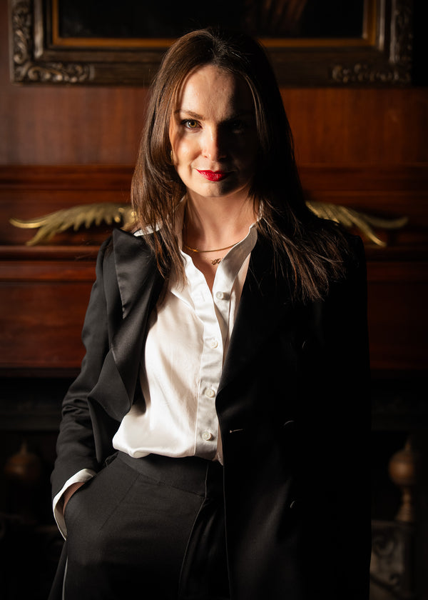 Woman in a black blazer and white shirt sitting in front of a wooden piano.