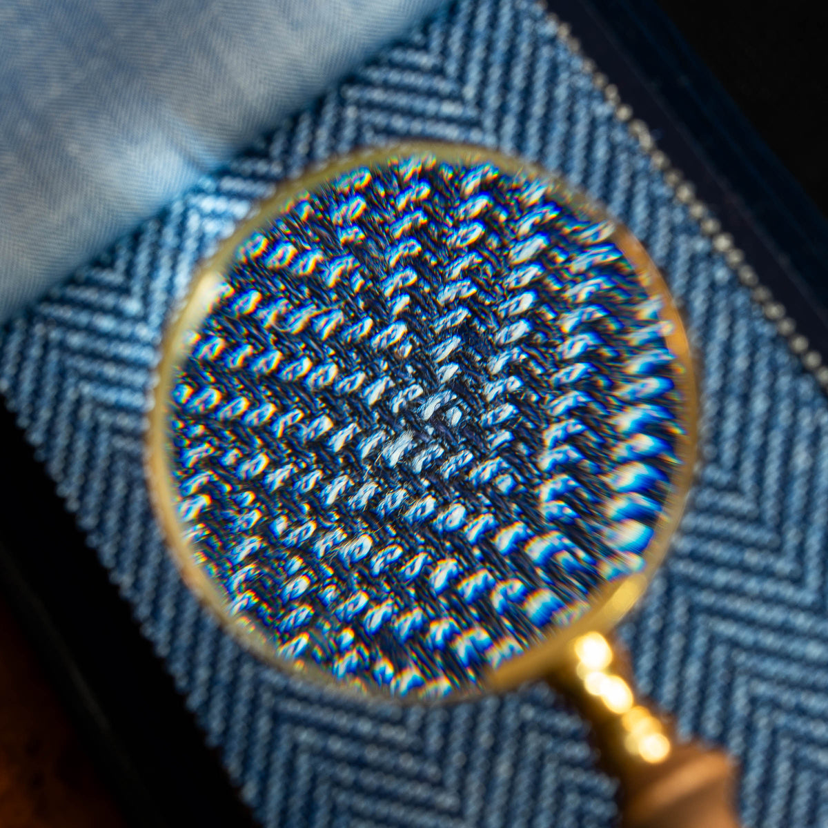 A close-up of a magnifying glass with a gold rim and wooden handle, showing a detailed view of a blue and white herringbone woven fabric texture.