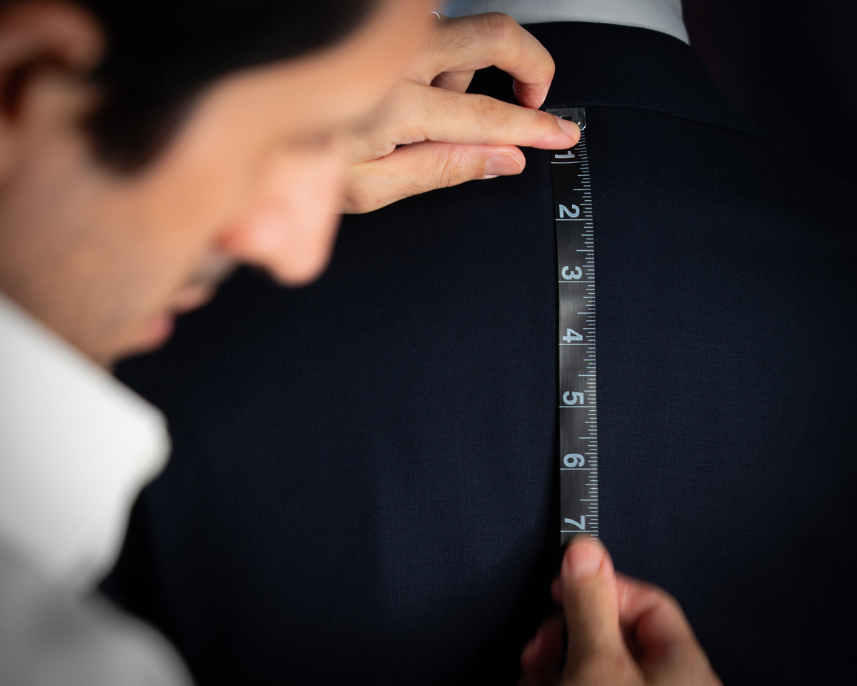A close-up shot of a tailor measuring the back of a dark navy suit jacket with a black and white tape measure, showing a person's hands and part of their face.