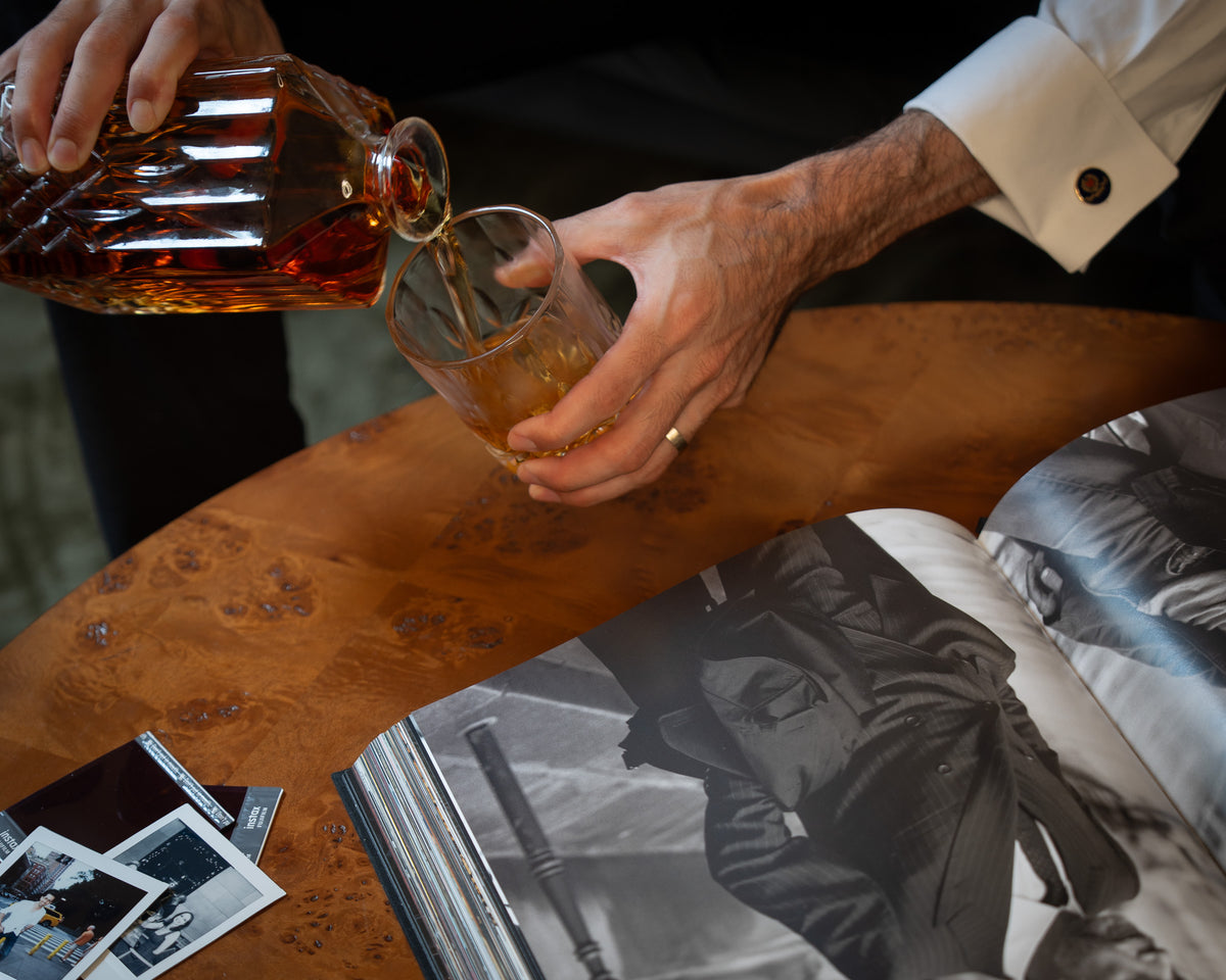 A close-up lifestyle shot of a person pouring amber liquor from a faceted glass decanter into a clear glass tumbler, set on a rustic wooden table with an open book and photos nearby.