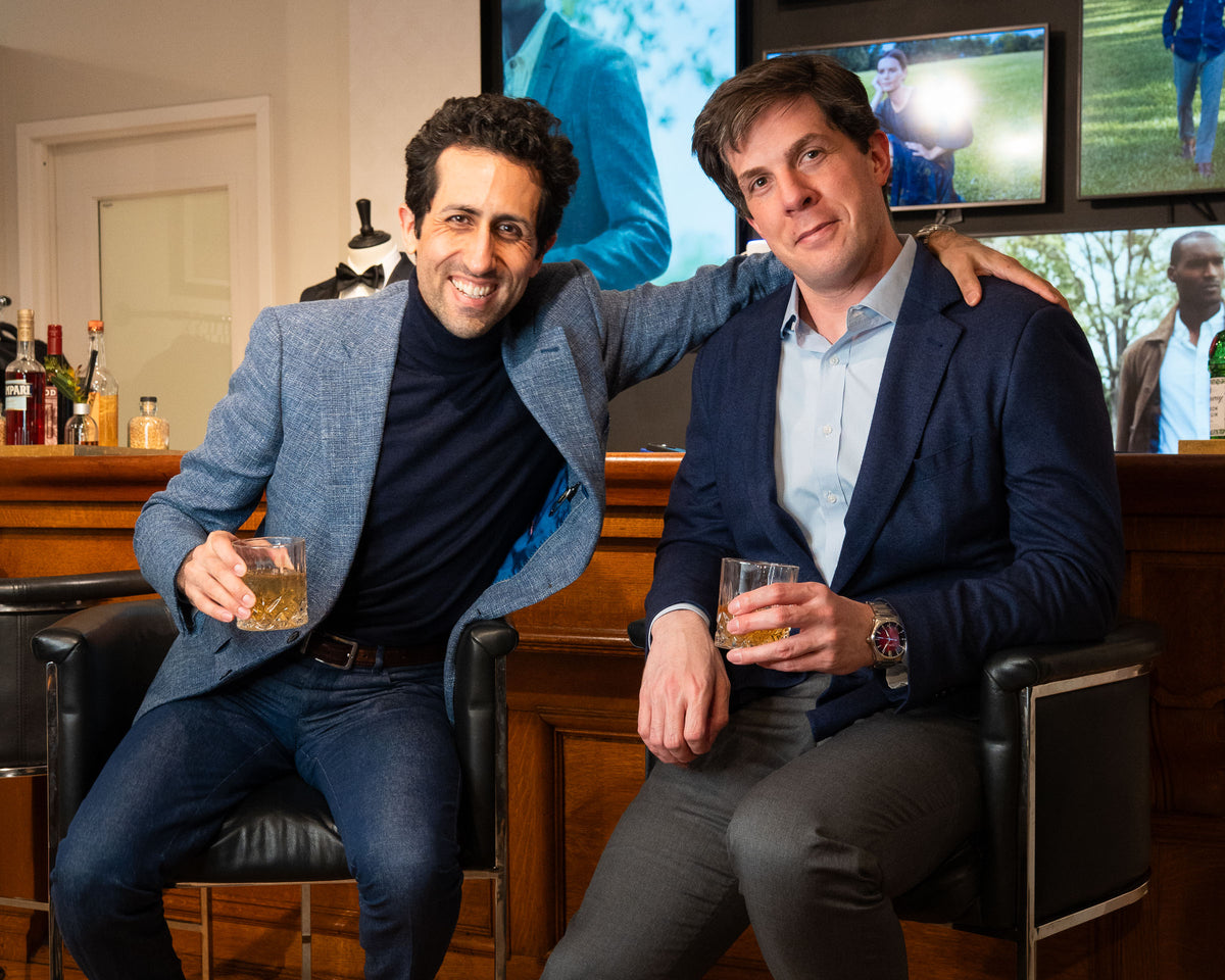 Two men sitting in chairs in a bar-like setting, each holding a glass of whiskey. The man on the left wears a blue blazer over a dark turtleneck and jeans, and the man on the right wears a navy blazer over a light blue dress shirt and gray trousers.