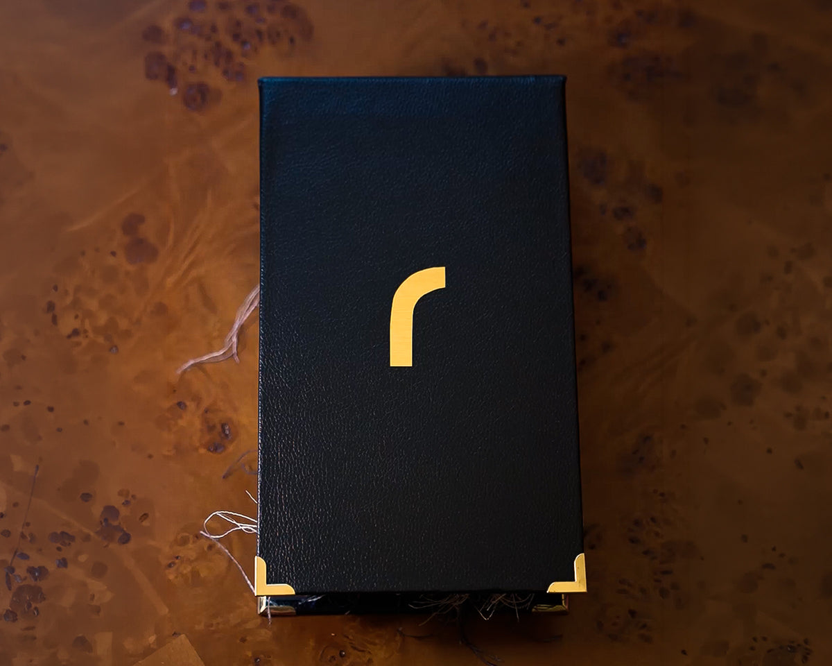 A black textured rectangular book with a gold stylized 'r' logo on the front and gold corner accents, photographed from above on a brown marbled surface.