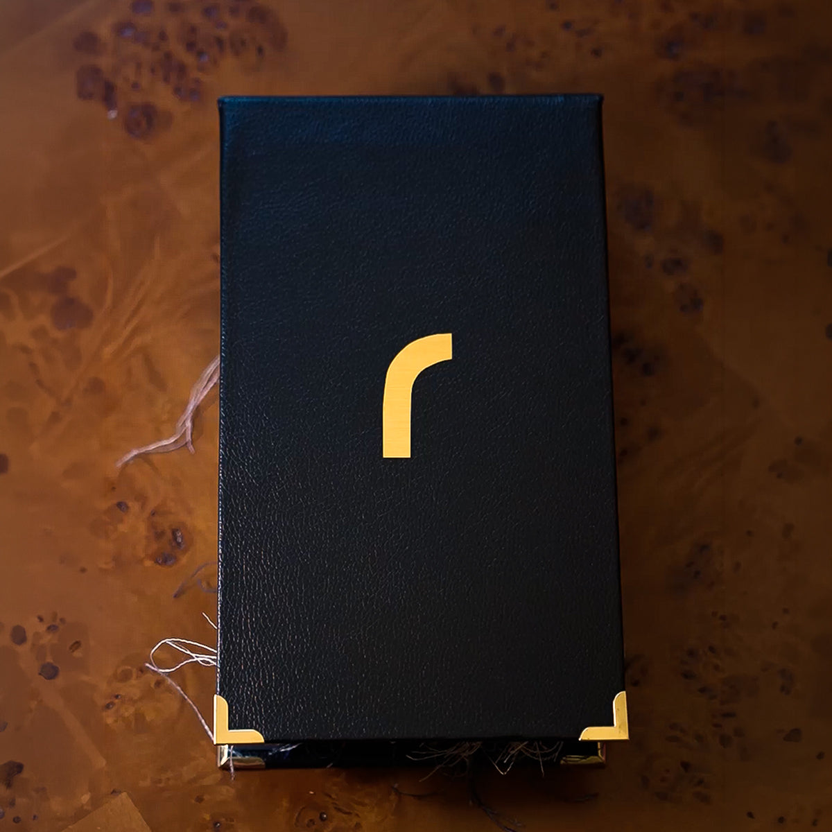 A black textured rectangular book with a gold stylized 'r' logo on the front and gold corner protectors, photographed from above on a brown marbled surface.