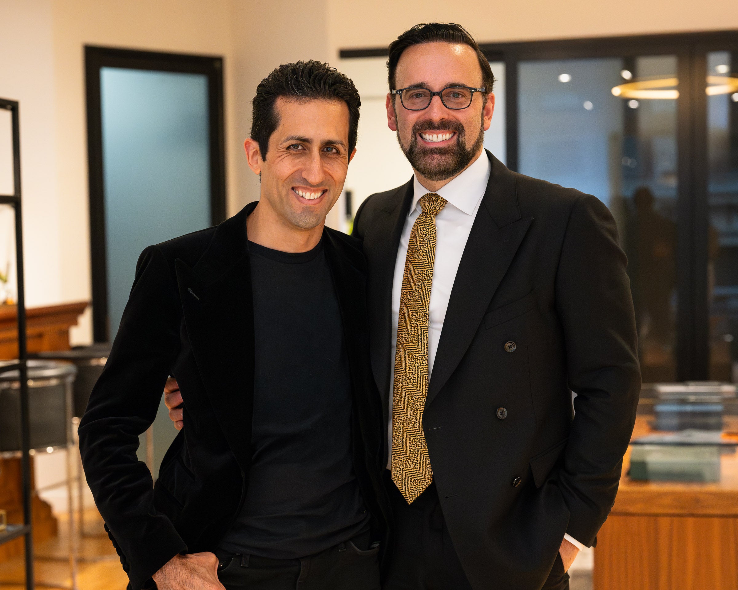 Two men standing together in an indoor office or showroom setting. The man on the left wears a black velvet blazer over a black shirt, and the man on the right wears a black double-breasted suit, white shirt, and a textured gold tie.