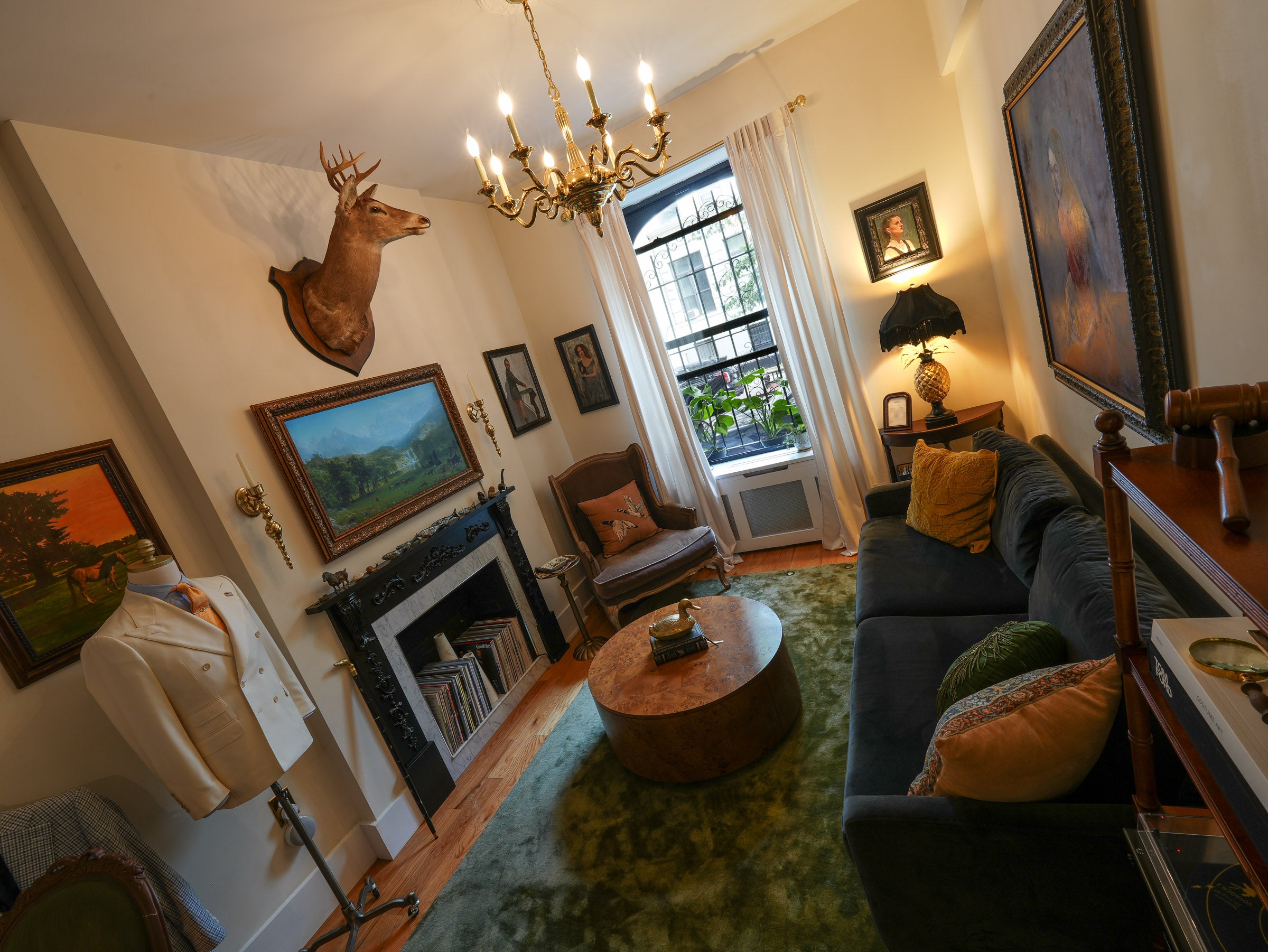 A vintage-style living room interior with a dark blue sofa, round wooden coffee table, green rug, fireplace, deer head wall mount, and multiple framed paintings.
