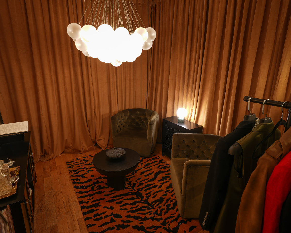 A cozy interior room with warm brown curtains, a glowing cloud-shaped pendant chandelier, a tiger-print rug, tufted armchairs, and a clothing rack with garments.