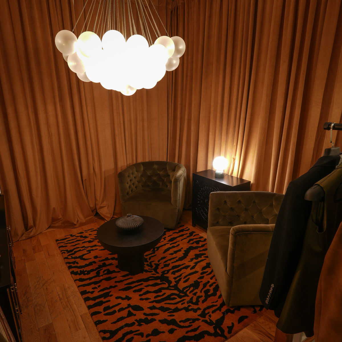 A cozy interior scene with warm brown curtains, a glowing cloud-style chandelier, two upholstered armchairs, a small round table, and a tiger-print rug on a wood floor.