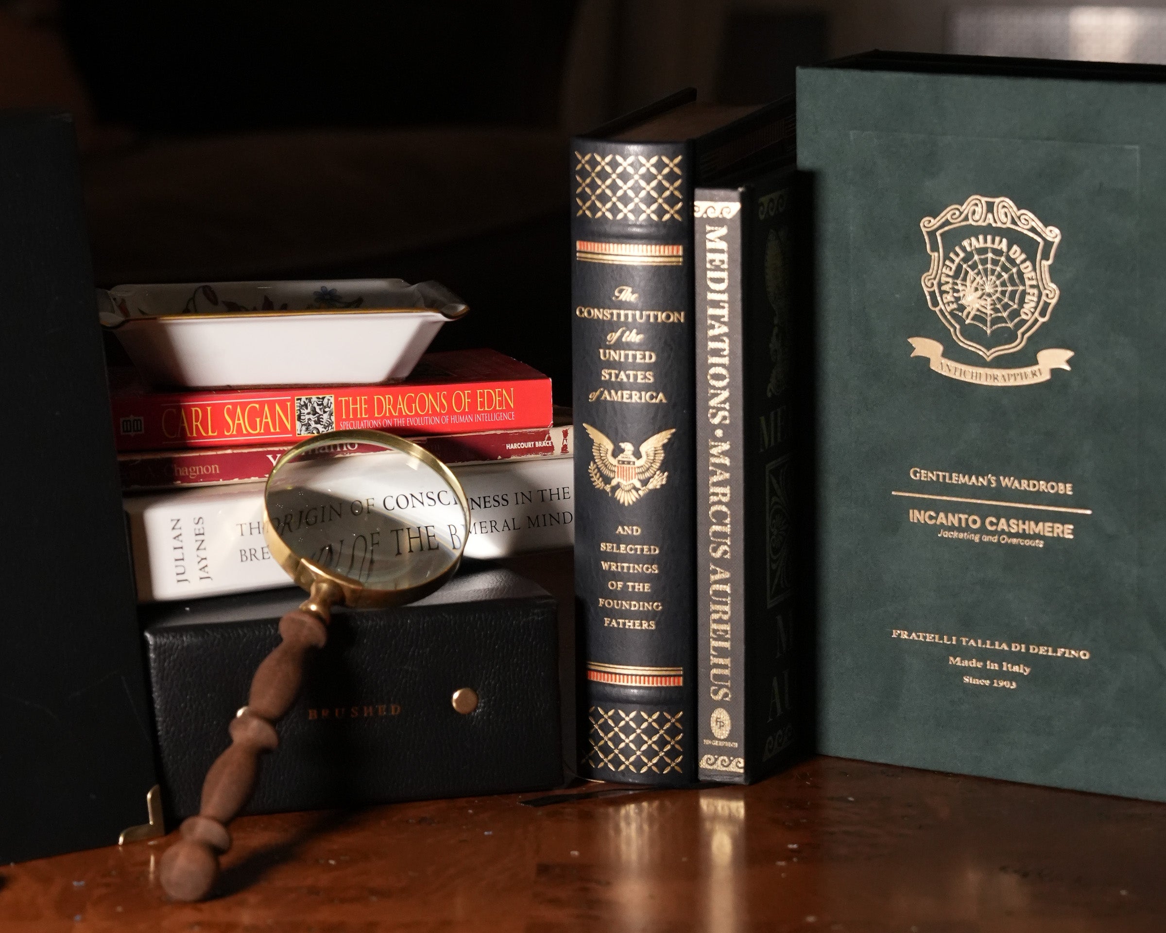 A dark green vintage-style book or journal standing upright on a wooden surface, next to three black books with gold lettering, a stack of books and a magnifying glass on a black box, and a dark background.