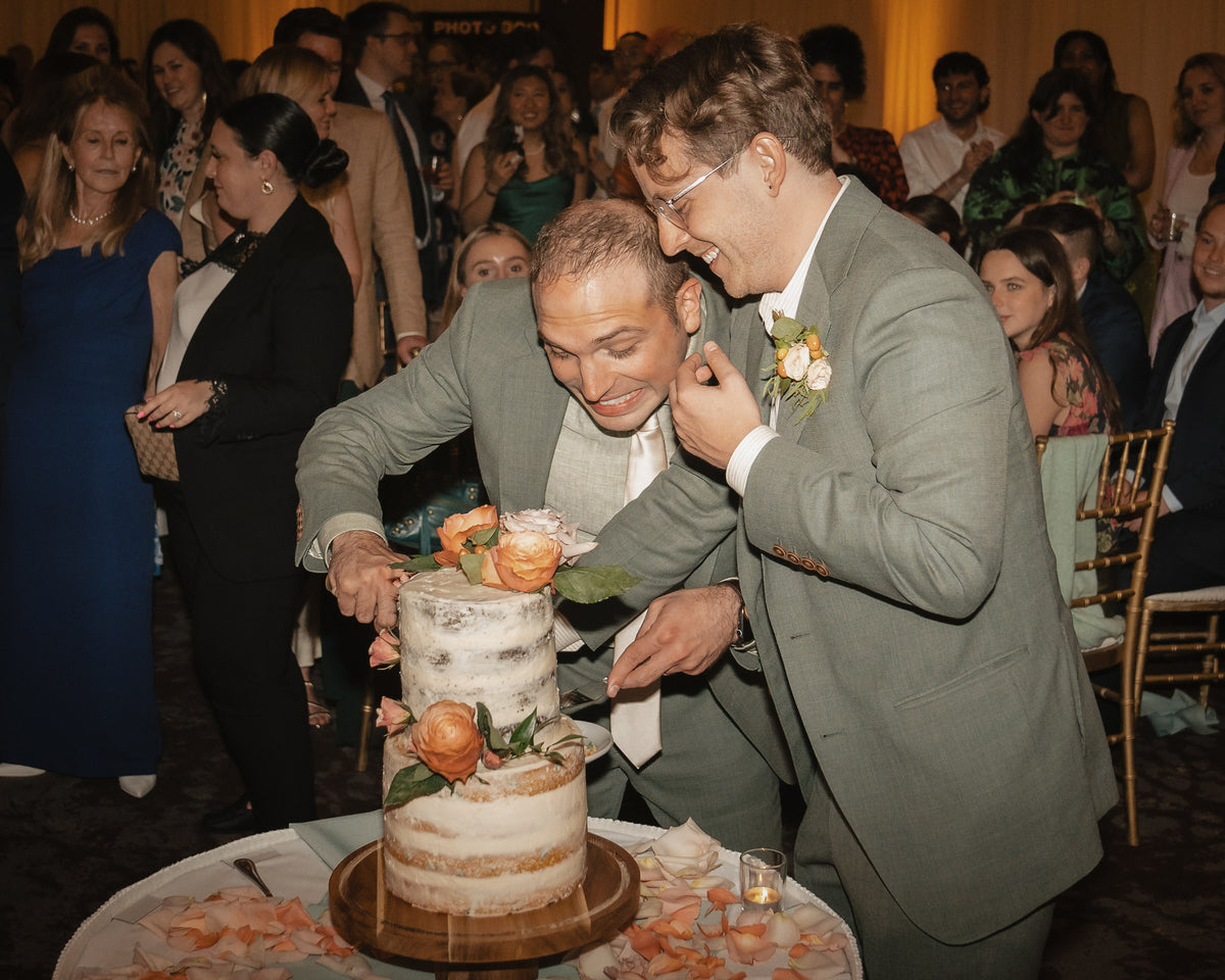 Two men in sage green suits cut into a two-tiered birch wood cake decorated with peach roses, surrounded by guests at an indoor reception.