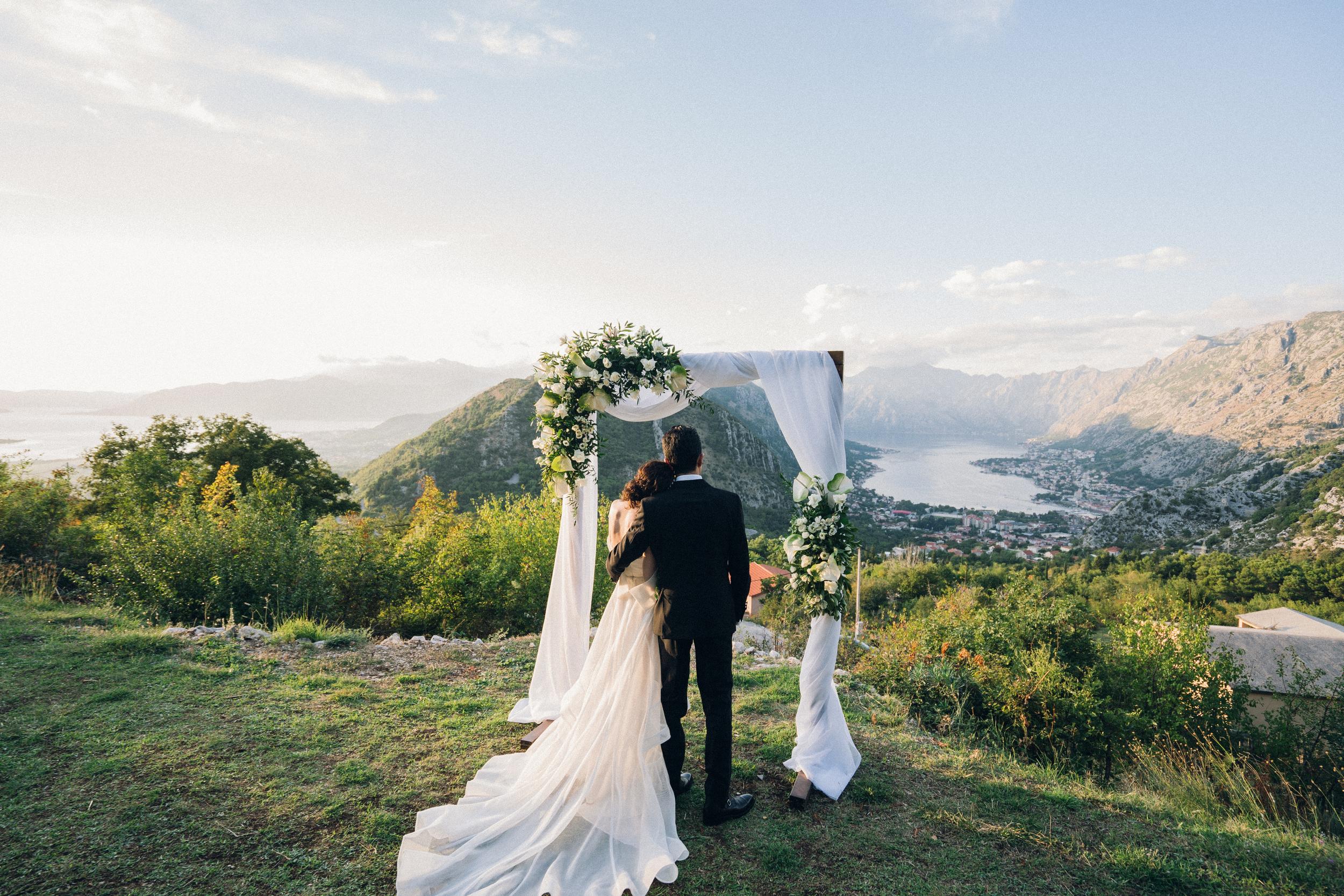 Couple standing under a wedding arch with a scenic view of mountains and water.